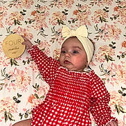 Gracie is registered to the contest to win money with this photo: baby, infant, girl, headband, bow, red_checkered_outfit, crib, floral_sheet, wooden_sign, four_months, milestone, cute, lying_down, hand, face, expression, indoor, child, person, young