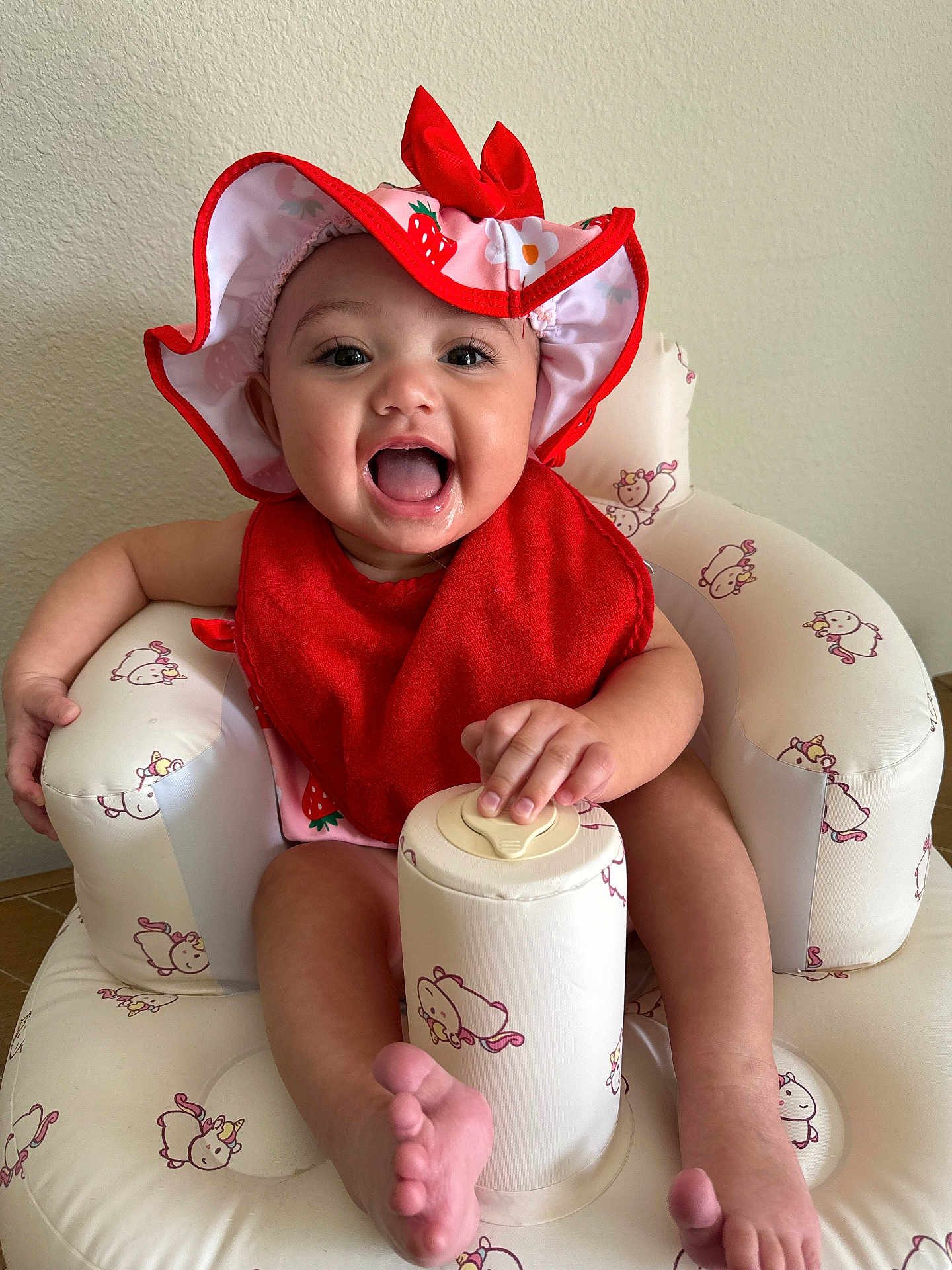 Airalee is registered to the contest to win money with this photo: baby, infant, smiling, hat, red_bib, unicorn_pattern, inflatable_chair, cute, happy, child, indoor, person, foot, hand, face, sitting, pink, red, playful, young
