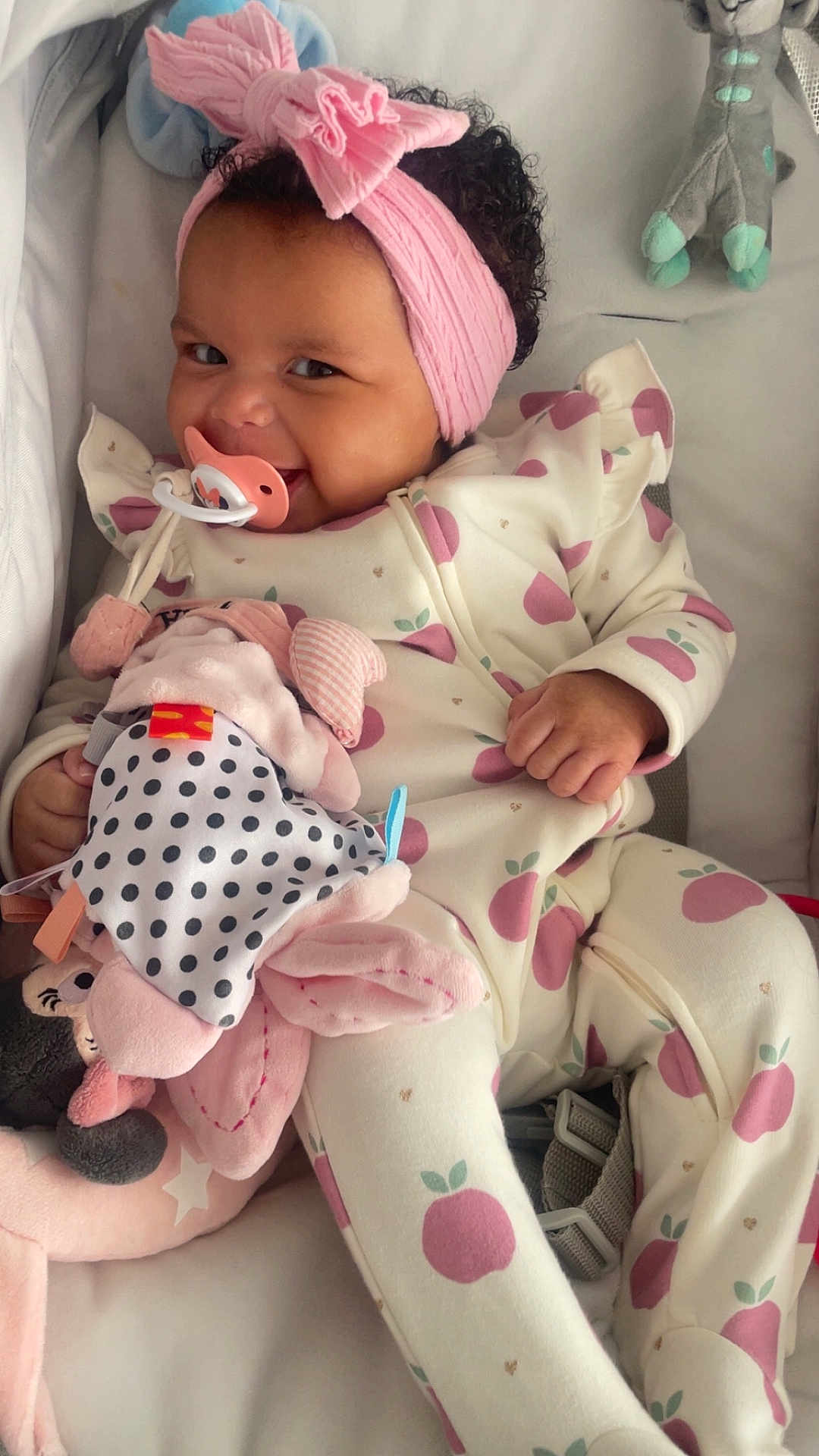 Naïla a rejoint le concours — aidez-le/la à gagner de superbes lots ! baby, infant, pacifier, headband, bow, onesie, apple_pattern, smiling, stuffed_animal, toy, curly_hair, chubby_cheeks, crib, blanket, pink, cozy, portrait, sitting, happy, nursery