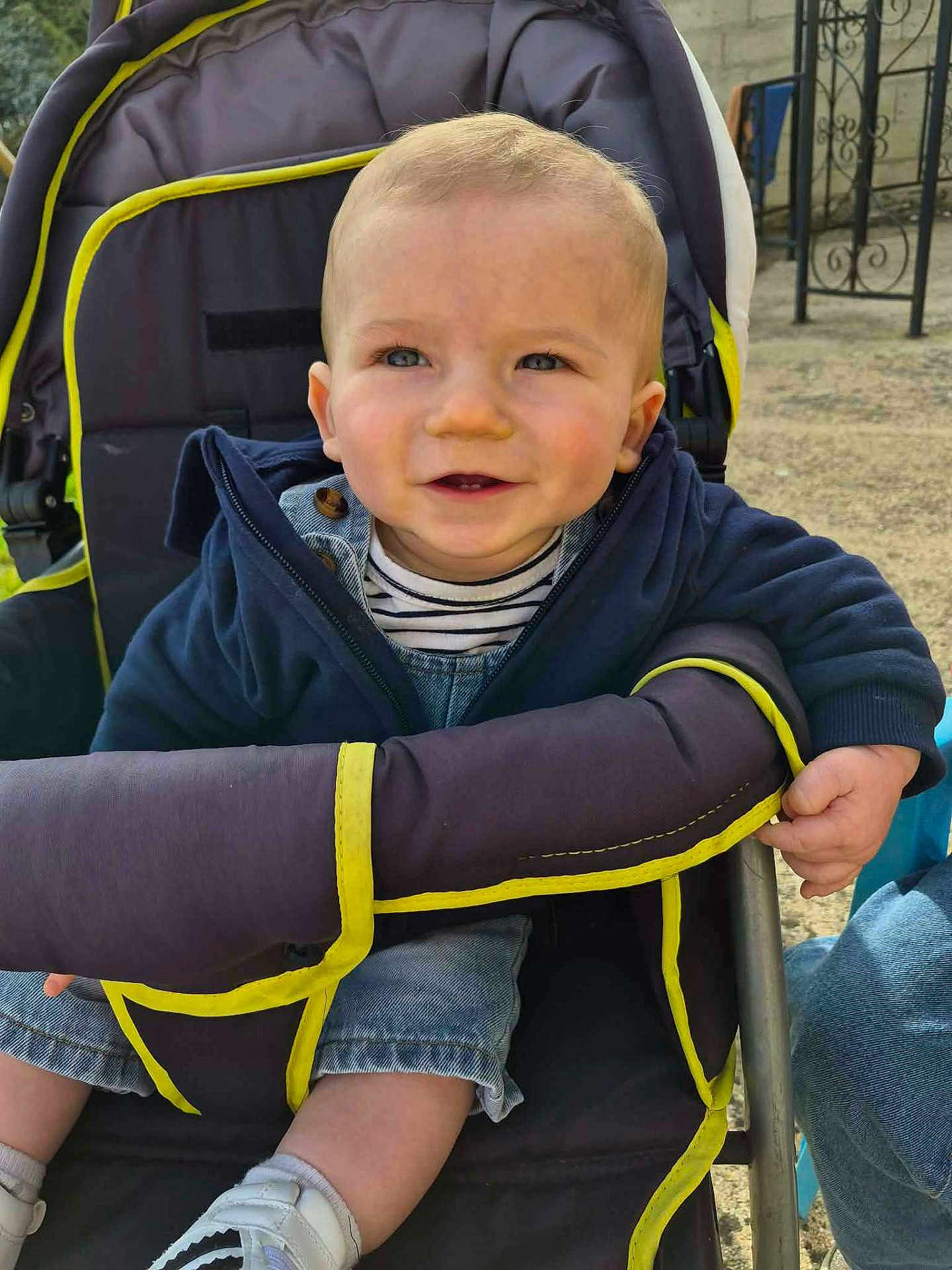 Maël participe au concours pour gagner de l'argent avec cette photo : baby, child, toddler, stroller, smile, portrait, outdoors, jacket, denim, striped_shirt, sneakers, hand, face, blue_eyes, cheeks, seat, pavement, fence, neon_trim, adult_jeans