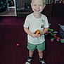 child, smiling, standing, toy, carpet, indoor, shorts, tshirt, sneakers, play, happy, hair, mohawk, room, furniture, storage, plastic_toys, floor, person, casual_clothing