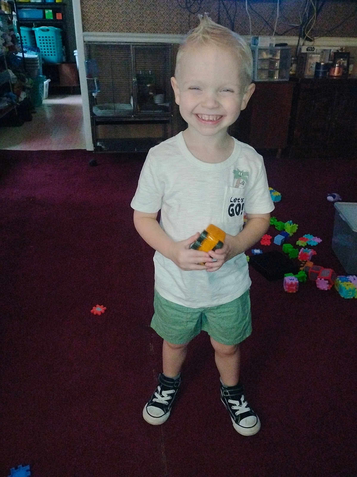 Westin Foote is registered to the contest to win money with this photo: child, smiling, standing, toy, carpet, indoor, shorts, tshirt, sneakers, play, happy, hair, mohawk, room, furniture, storage, plastic_toys, floor, person, casual_clothing