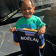 Noëlan participe au concours pour gagner de l'argent avec cette photo : child, boy, smiling, braided_hair, green_tshirt, spider_man, holding_shirt, navy_shirt, name_printed, indoor, floor_tiles, sunlight, shadow, happy, person, portrait, casual_clothing, young_child, standing, bright_light