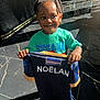 child, boy, smiling, braided_hair, green_tshirt, spider_man, holding_shirt, navy_shirt, name_printed, indoor, floor_tiles, sunlight, shadow, happy, person, portrait, casual_clothing, young_child, standing, bright_light
