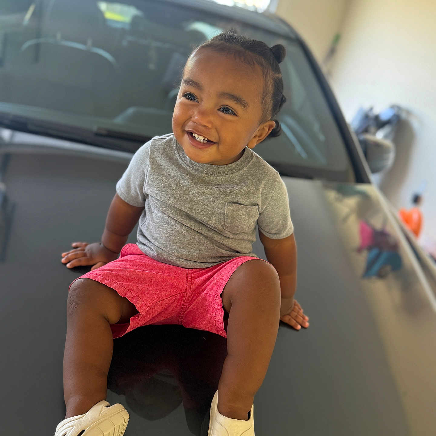 Liam joined the competition — help win amazing prizes! car, casual_clothing, child, cute, footwear, garage, gray_shirt, hair_buns, happy, hood, indoor, person, pink_shorts, portrait, reflection, sitting, smiling, toddler, white_shoes, young_child
