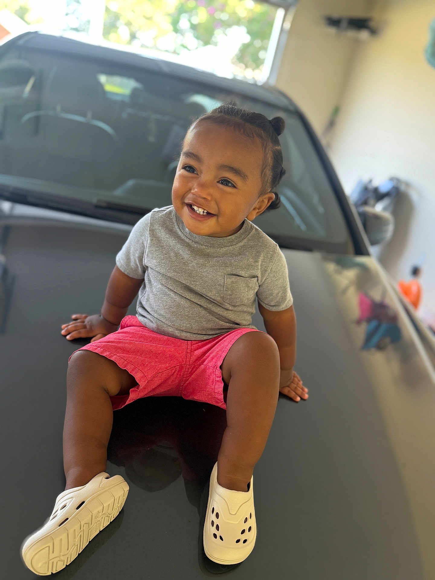 Liam joined the competition — help win amazing prizes! toddler, child, smiling, happy, sitting, car, hood, gray_shirt, pink_shorts, white_shoes, indoor, garage, reflection, cute, person, portrait, hair_buns, footwear, casual_clothing, young_child