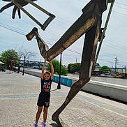 Paola is registered to the contest to win money with this photo: child, sunglasses, outdoor, sculpture, metal, statue, river, bridge, pavement, daylight, shadow, trees, lamp_post, playful, urban, colorful_clothing, person, summer, happy, public_space