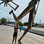 child, sunglasses, outdoor, sculpture, metal, statue, river, bridge, pavement, daylight, shadow, trees, lamp_post, playful, urban, colorful_clothing, person, summer, happy, public_space