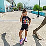 child, girl, sunglasses, posing, outdoor, sidewalk, bridge, river, street_lamp, shadow, casual_clothing, shorts, tshirt, sneakers, urban, daylight, tree, metal_structure, smiling, fashion