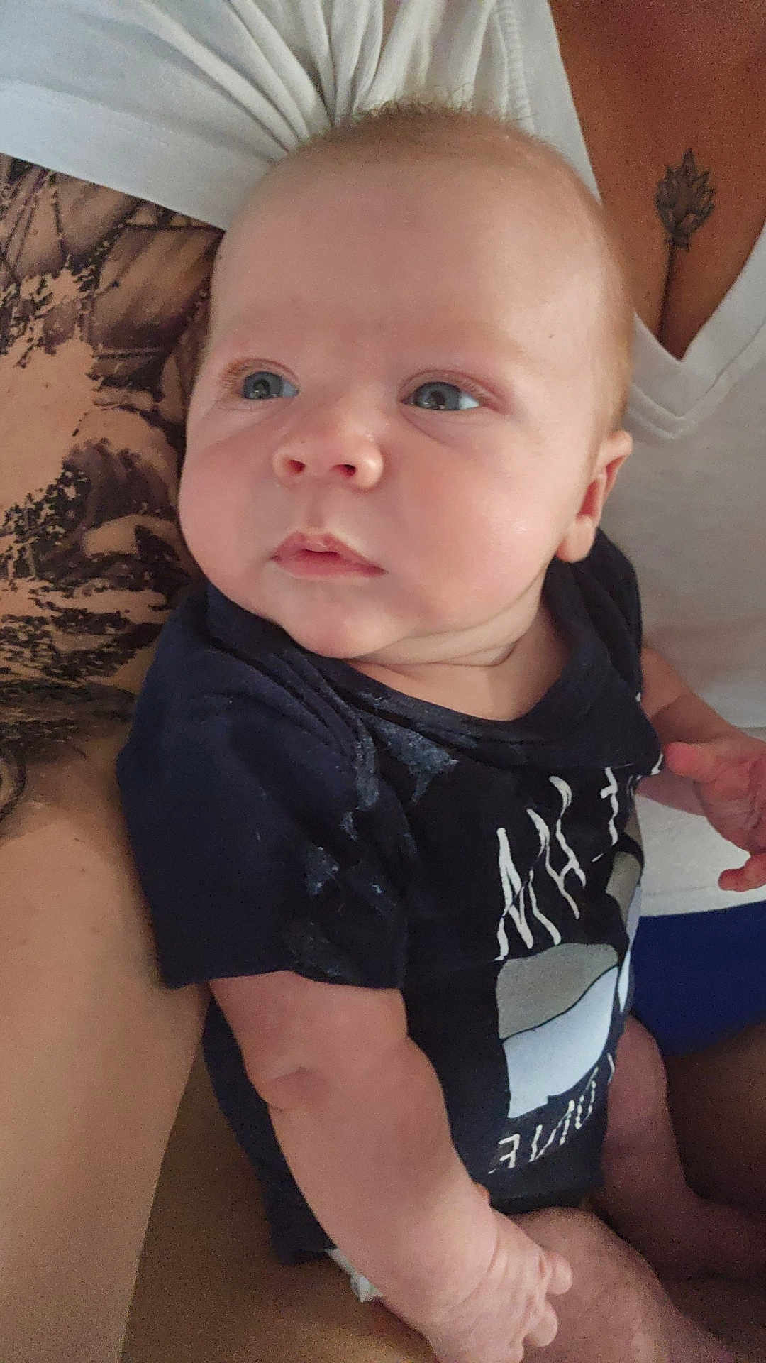 Curtis is registered to the contest to win money with this photo: baby, infant, person, arm, tattoo, shirt, clothing, holding, skin, face, portrait, child, human, closeup, casual, indoor, cute, newborn, expression, family