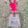 child, girl, barefoot, red_dress, flowers, grave, gravestone, grass, outdoor, smiling, person, adult, holding, memorial, headstone, cemetery, nature, happy, casual_clothing, supporting_hand