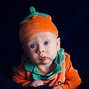 Aryton is registered to the contest to win money with this photo: baby, infant, child, pumpkin_costume, orange_hat, blue_eyes, cute, portrait, costume, soft_lighting, dark_background, curious_expression, hands, face, headwear, fall_theme, seasonal, closeup, clothing, adorable