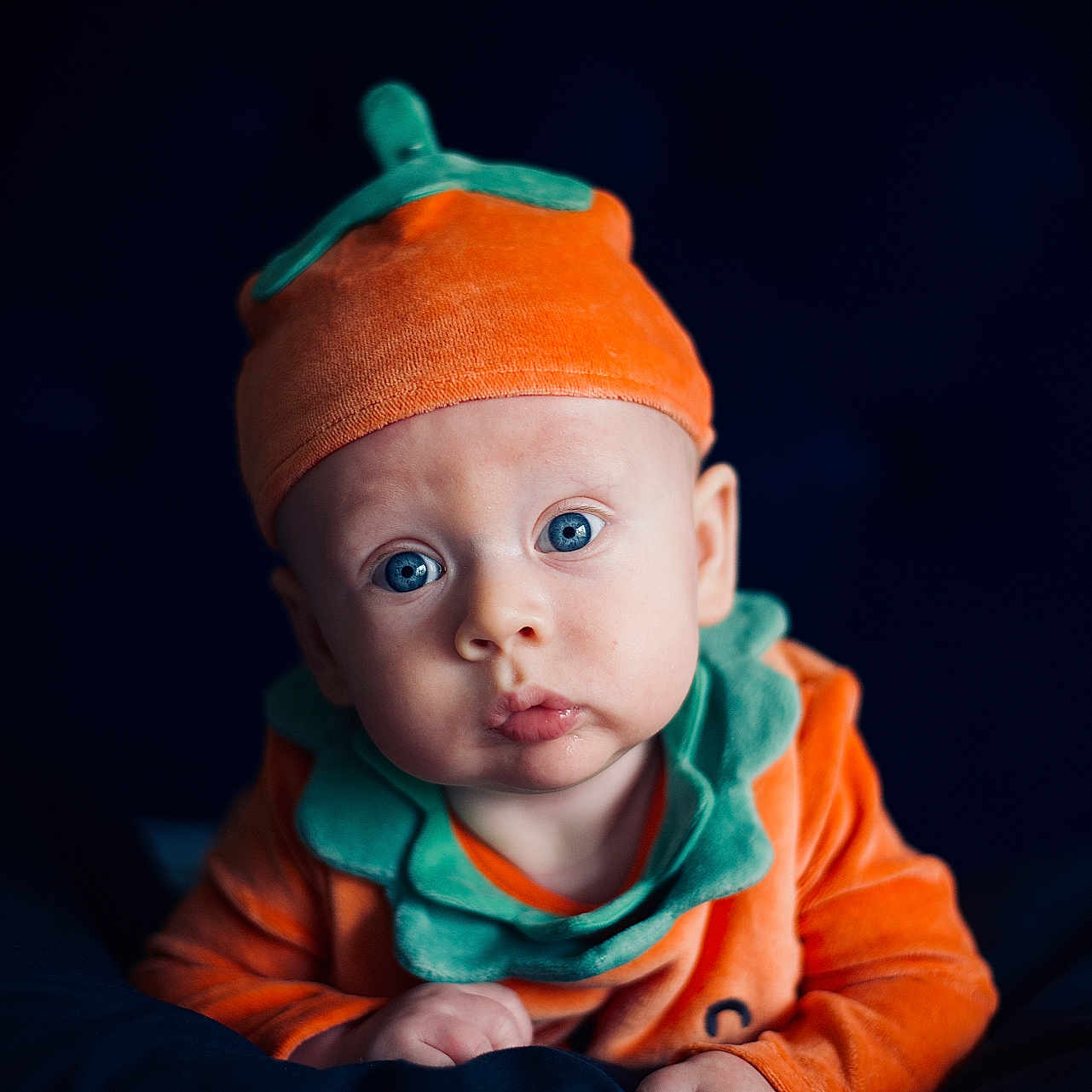 Aryton is registered to the contest to win money with this photo: adorable, baby, blue_eyes, child, closeup, clothing, costume, curious_expression, cute, dark_background, face, fall_theme, hands, headwear, infant, orange_hat, portrait, pumpkin_costume, seasonal, soft_lighting