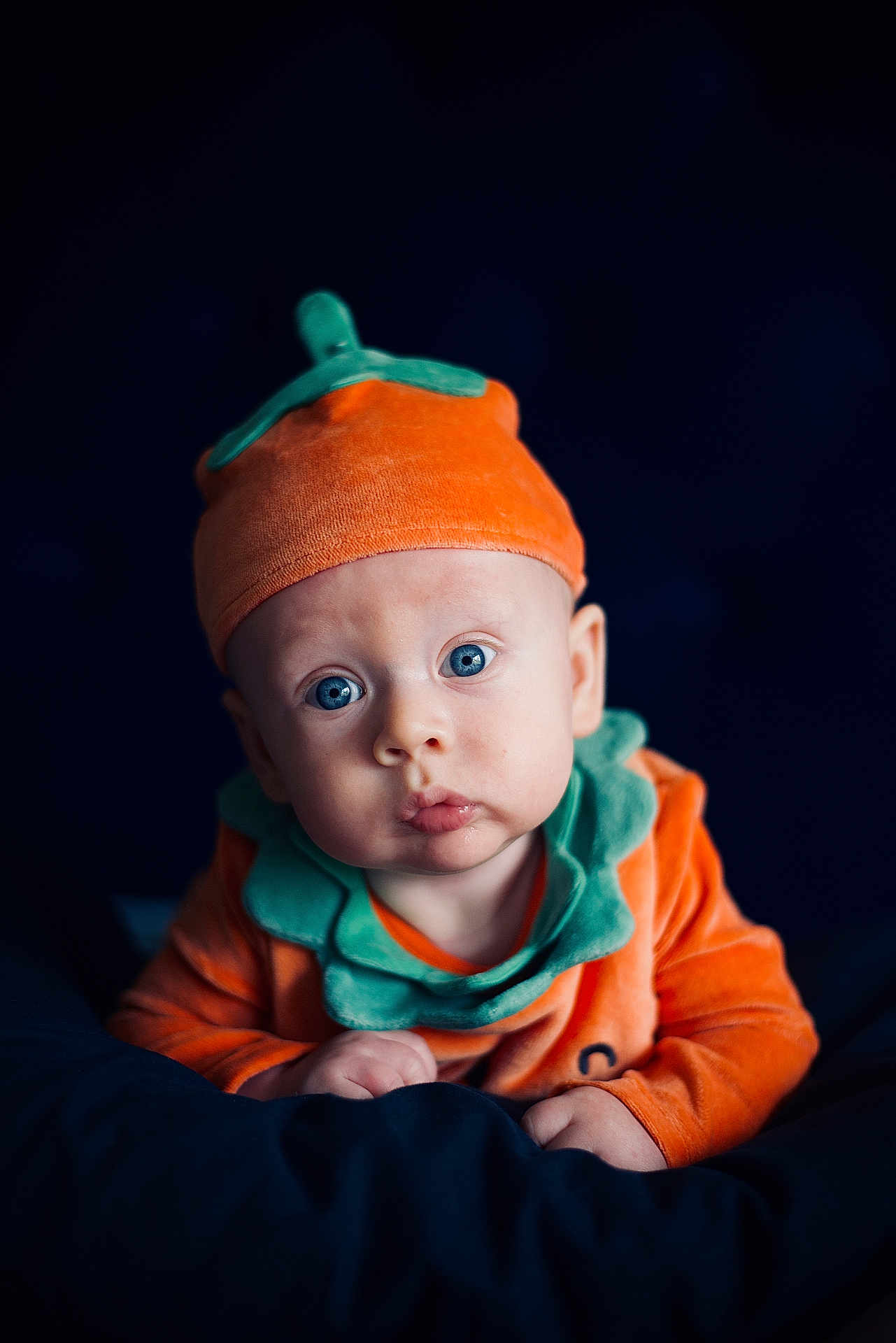 Aryton is registered to the contest to win money with this photo: baby, infant, child, pumpkin_costume, orange_hat, blue_eyes, cute, portrait, costume, soft_lighting, dark_background, curious_expression, hands, face, headwear, fall_theme, seasonal, closeup, clothing, adorable