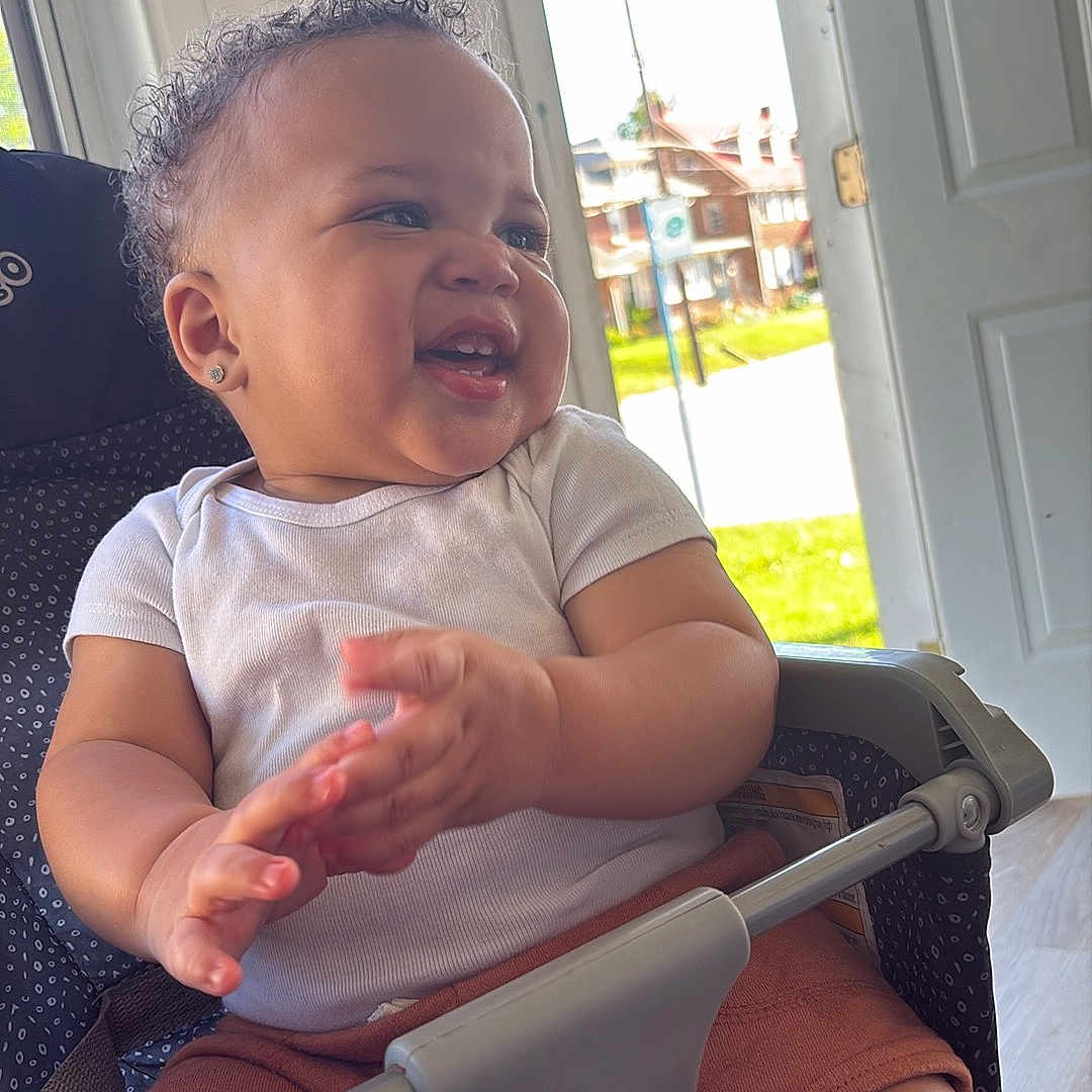 Xykie is registered to the contest to win money with this photo: baby, brown_shorts, child, clapping, curly_hair, daylight, door, earrings, expression, footwear, happy, indoors, infant, person, seatbelt, skin, smiling, stroller, white_shirt, window