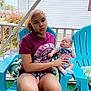 woman, baby, child, outdoor, porch, plastic_chair, turquoise, tshirt, shorts, tattoo, curly_hair, baby_clothes, greenery, cushion, fence, house, daylight, expression, holding, pout