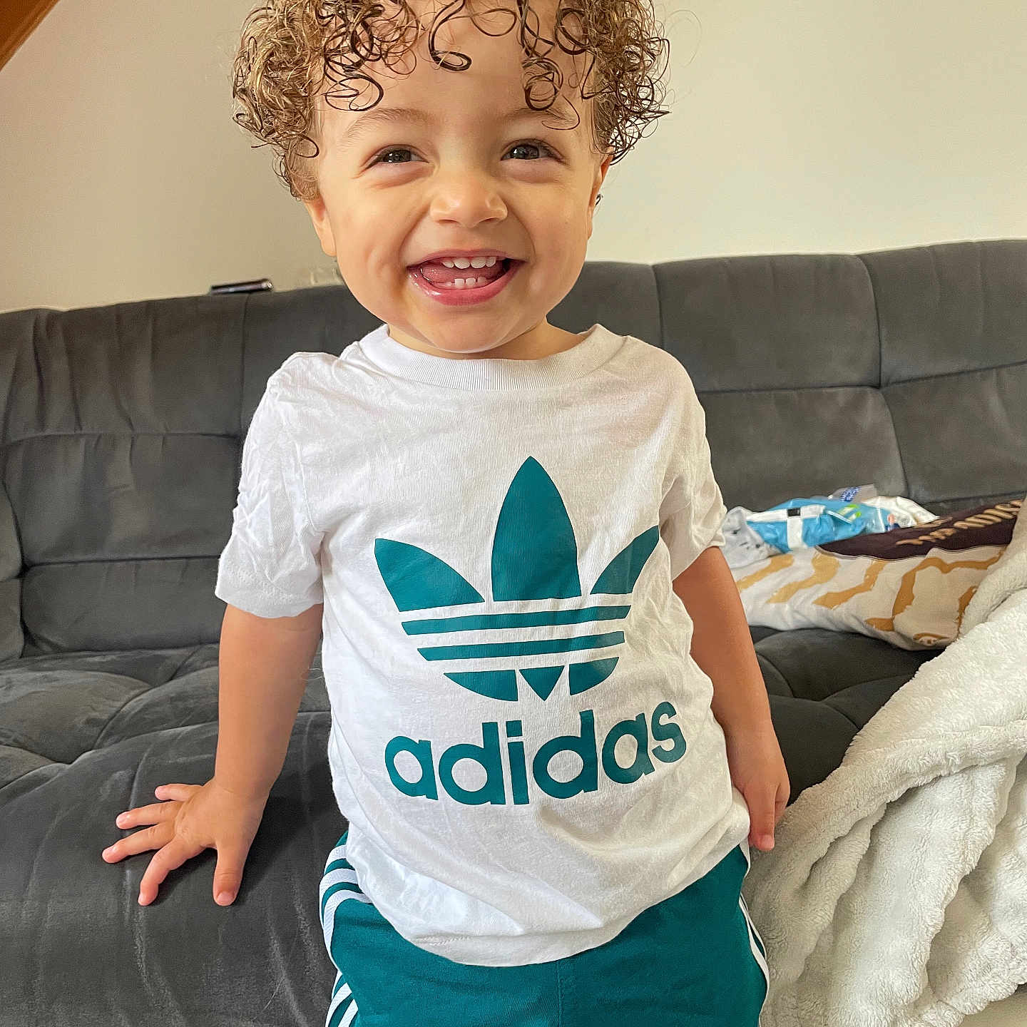 Keymïs participe au concours pour gagner de l'argent avec cette photo : adidas_logo, blanket, casual, child, clothing, couch, curly_hair, cute, furniture, green_shorts, happy, indoor, living_room, person, portrait, smiling, standing, toddler, white_tshirt, young_child