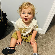 Denver is registered to the contest to win money with this photo: toddler, child, curly_hair, blue_eyes, smiling, sitting, carpet, indoor, beige_shirt, nike_logo, shorts, sandals, footwear, wall, baseboard, hand, happy, young_child, person, casual_clothing