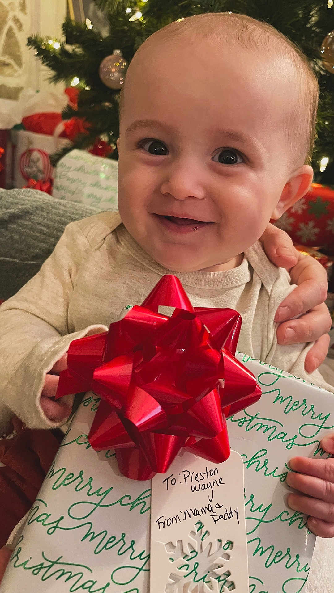 Preston is registered to the contest to win money with this photo: baby, child, smiling, present, gift, red_bow, christmas, holiday, tree, decorations, indoor, cute, happy, person, festive, celebration, wrapped_gift, hand, tag, cozy