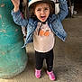 child, helmet, denim_jacket, pink_shoes, black_pants, concrete_floor, green_pillar, barn, playful, excited, standing, curly_hair, indoor, casual_clothing, person, smiling, happy, rustic, farm_tools, expression