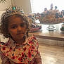 child, girl, tiara, crown, dress, floral, red_skirt, curly_hair, indoor, cake, dolls, table, flooring, window, natural_light, party, celebration, food, birthday, portrait