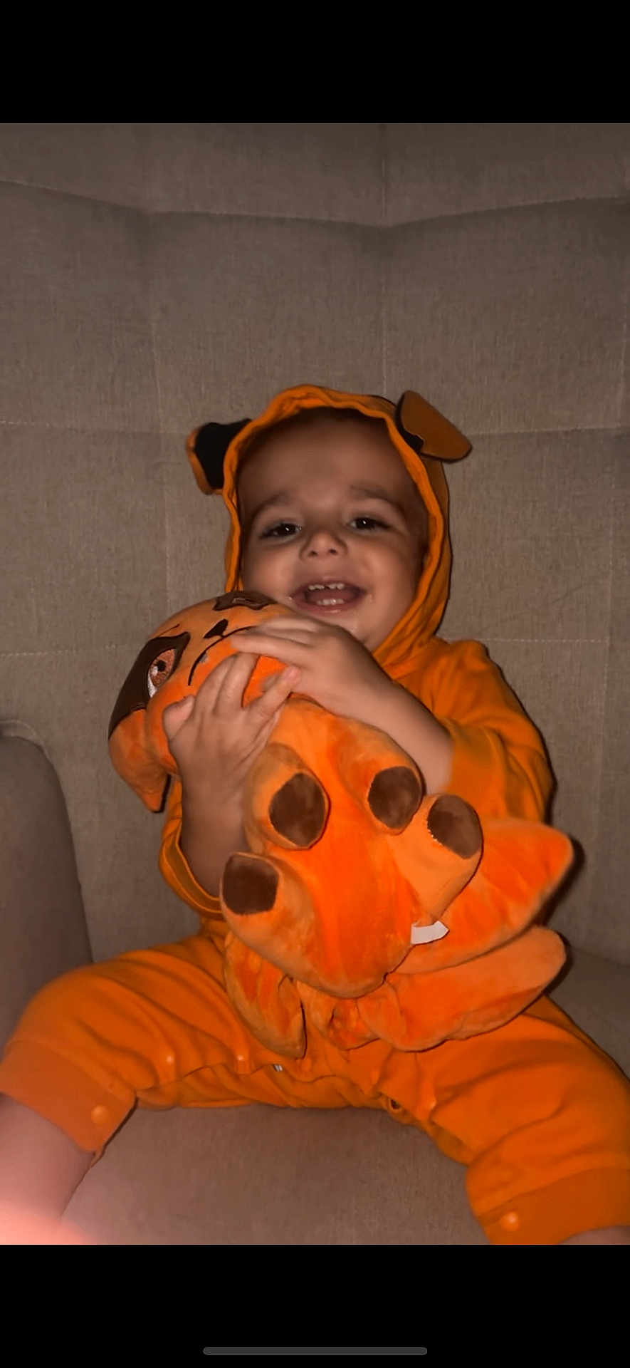 Aurélien participe au concours pour gagner de l'argent avec cette photo : toddler, child, plush_toy, orange_onesie, dog_costume, smiling, happy, couch, indoor, cozy, cute, soft_toy, childrens_clothing, seated, playful, infant, baby, fun, warm_colors, portrait