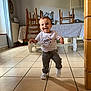Aurélien participe au concours pour gagner de l'argent avec cette photo : toddler, child, smiling, indoor, kitchen, table, chairs, tile_floor, natural_light, casual_clothing, white_shirt, pants, sneakers, radiator, curtains, wooden_furniture, home, person, baby, walking