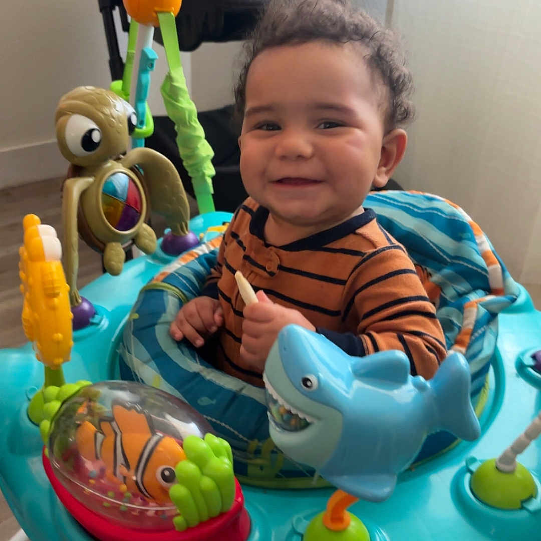 Kobe is registered to the contest to win money with this photo: toddler, child, smile, activity_center, toy_shark, toy_fish, toy_turtle, indoor, baby, playtime, striped_clothing, curly_hair, flooring, window_light, happy, seat, colorful, plastic_toys, baby_activity, infant