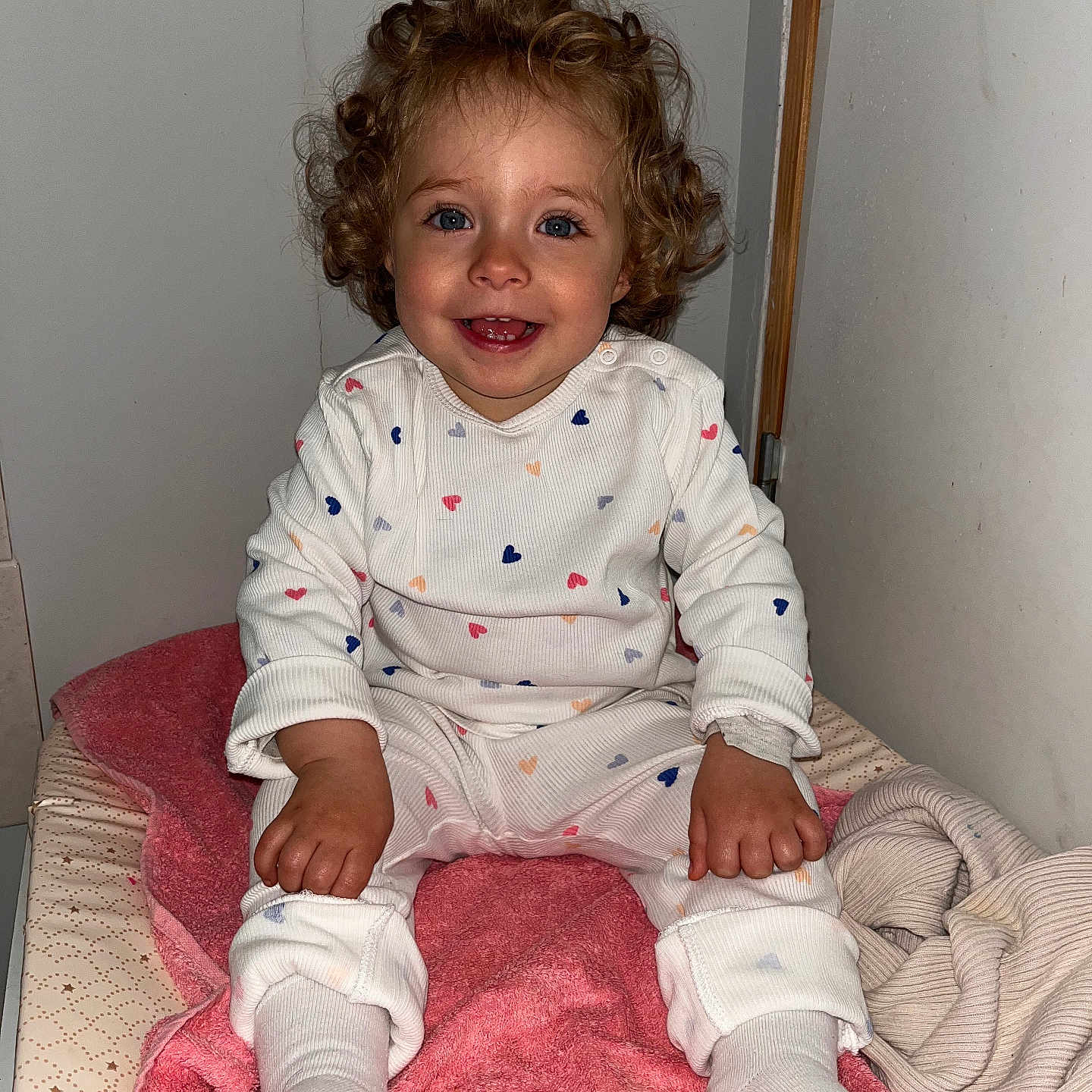 Charlotte participe au concours pour gagner de l'argent avec cette photo : baby, blanket, blue_eyes, child, clothing, cozy, curly_hair, cute, feet, hands, happy, hearts_pattern, indoors, person, pink_towel, portrait, sitting, smiling, toddler, white_outfit