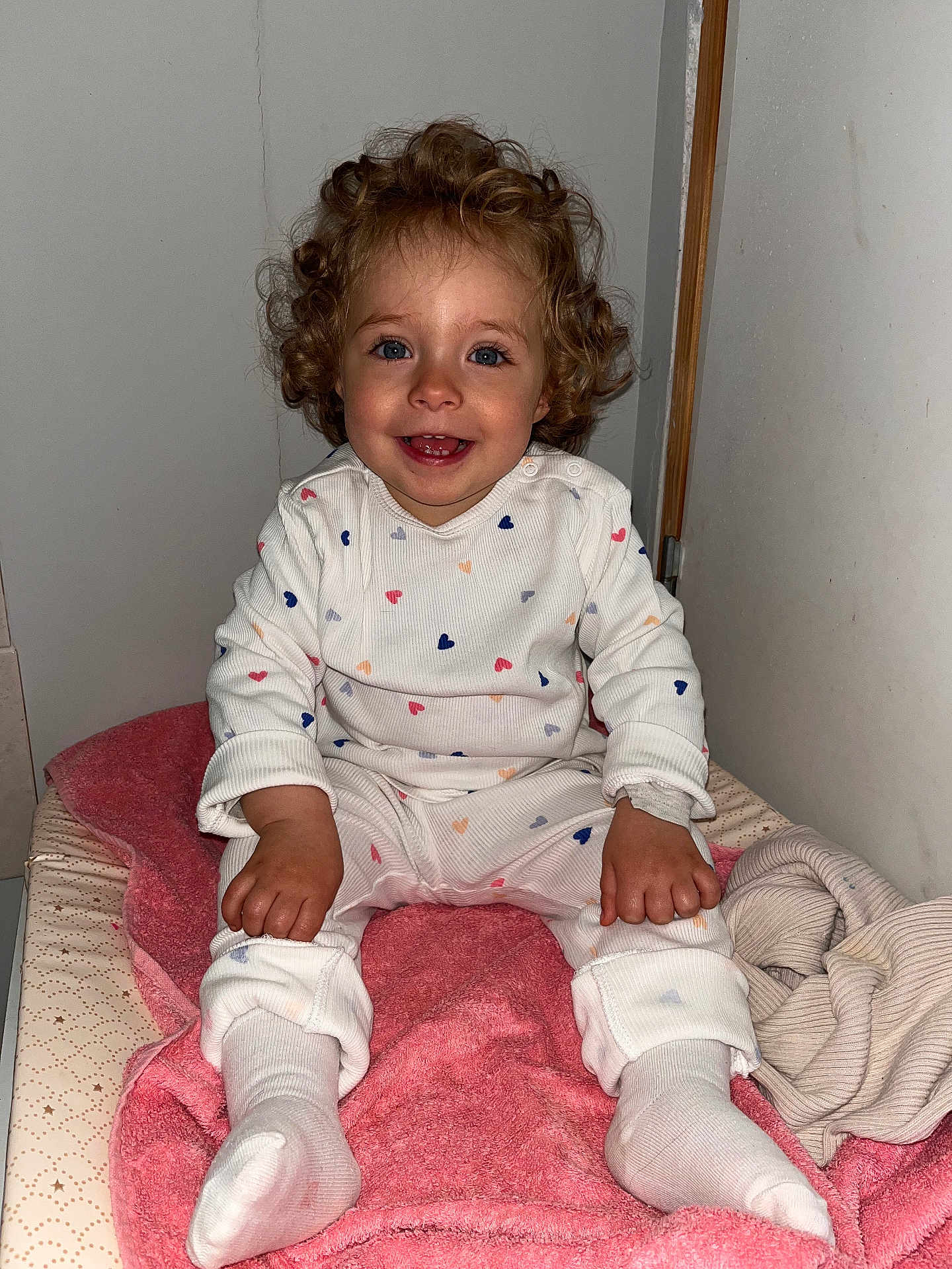 Charlotte participe au concours pour gagner de l'argent avec cette photo : toddler, child, curly_hair, blue_eyes, white_outfit, hearts_pattern, pink_towel, sitting, indoors, smiling, happy, cozy, blanket, clothing, person, portrait, cute, baby, feet, hands