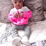 D'Aruia is registered to the contest to win money with this photo: baby, child, infant, pink_jacket, headband, sitting, bed, pillow, blanket, clothing, person, indoor, portrait, cute, white_shoes, gray_pants, face, young_child, soft_texture, curious_expression