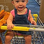 child, toddler, shopping_cart, denim_overalls, pink_bow, pink_shoes, retail_store, aisle, blue_floor, hand, smile, face, person, clothing, bag, yellow_handle, indoor, cute, baby, footwear