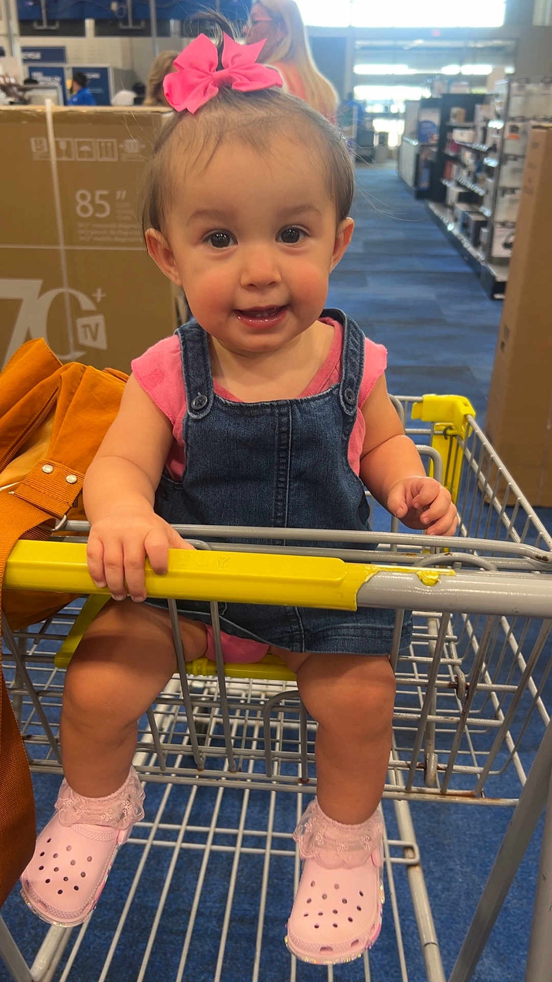 Aliviah joined the competition — help win amazing prizes! child, toddler, shopping_cart, denim_overalls, pink_bow, pink_shoes, retail_store, aisle, blue_floor, hand, smile, face, person, clothing, bag, yellow_handle, indoor, cute, baby, footwear