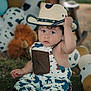 baby, balloons, child, costume, cow_pattern, cowboy_hat, curly_hair, cute, expression, foot, grass, hat, outdoor, overalls, playful, portrait, sitting, stuffed_animal, toddler, toy