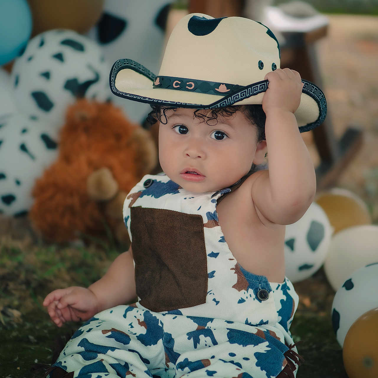 Liam joined the competition — help win amazing prizes! baby, balloons, child, costume, cow_pattern, cowboy_hat, curly_hair, cute, expression, foot, grass, hat, outdoor, overalls, playful, portrait, sitting, stuffed_animal, toddler, toy