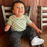 Liam is registered to the contest to win money with this photo: toddler, child, smiling, curly_hair, checkered_shirt, black_pants, white_sneakers, wooden_chair, metal_chair, wooden_wall, indoor, happy, sitting, cute, portrait, person, fashion, casual, looking_up, young_child