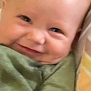 Shane is registered to the contest to win money with this photo: baby, infant, smile, face, eyes, cheeks, bald_head, onesie, green_clothing, pillow, blanket, patterned_background, close_up, portrait, indoors, cuteness, newborn, skin, cozy, grinning
