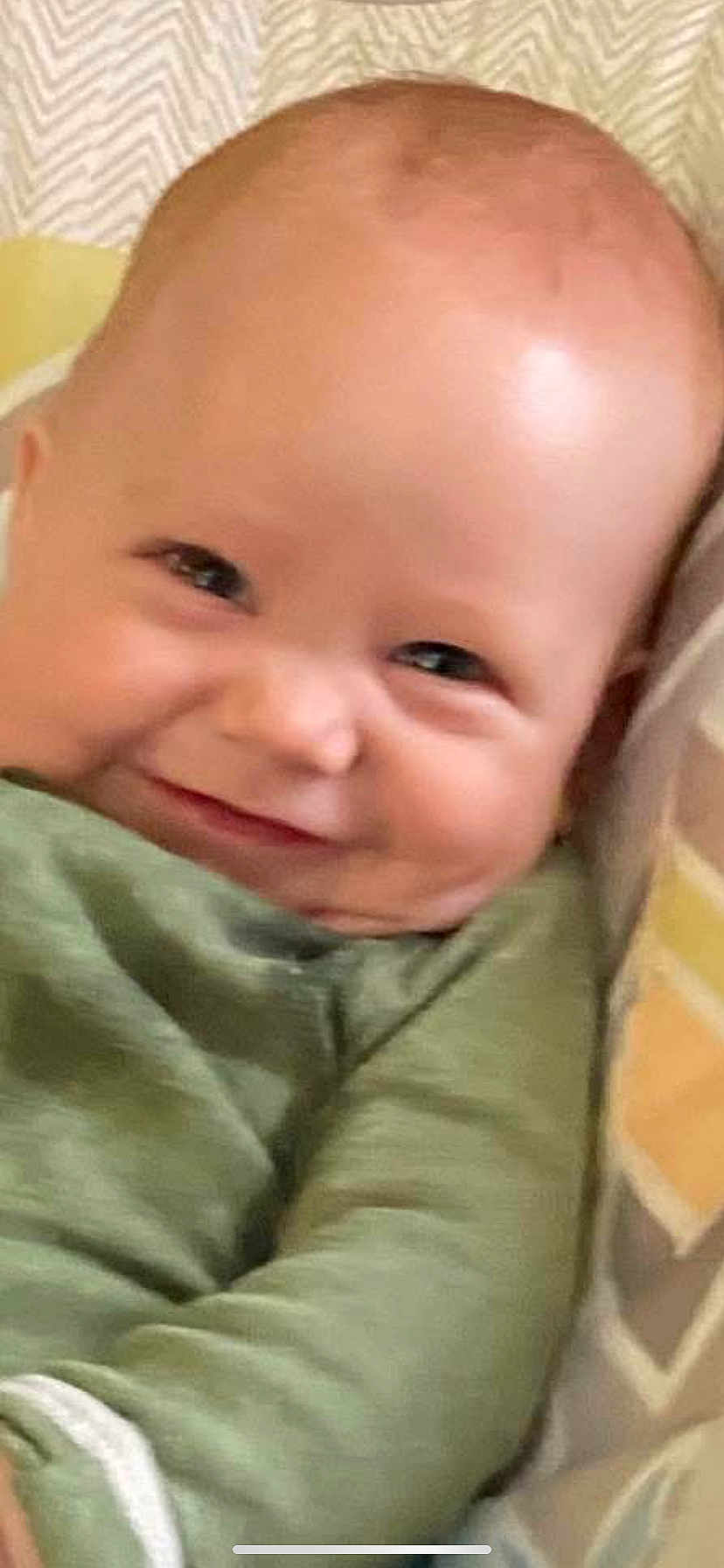 Shane is registered to the contest to win money with this photo: baby, infant, smile, face, eyes, cheeks, bald_head, onesie, green_clothing, pillow, blanket, patterned_background, close_up, portrait, indoors, cuteness, newborn, skin, cozy, grinning