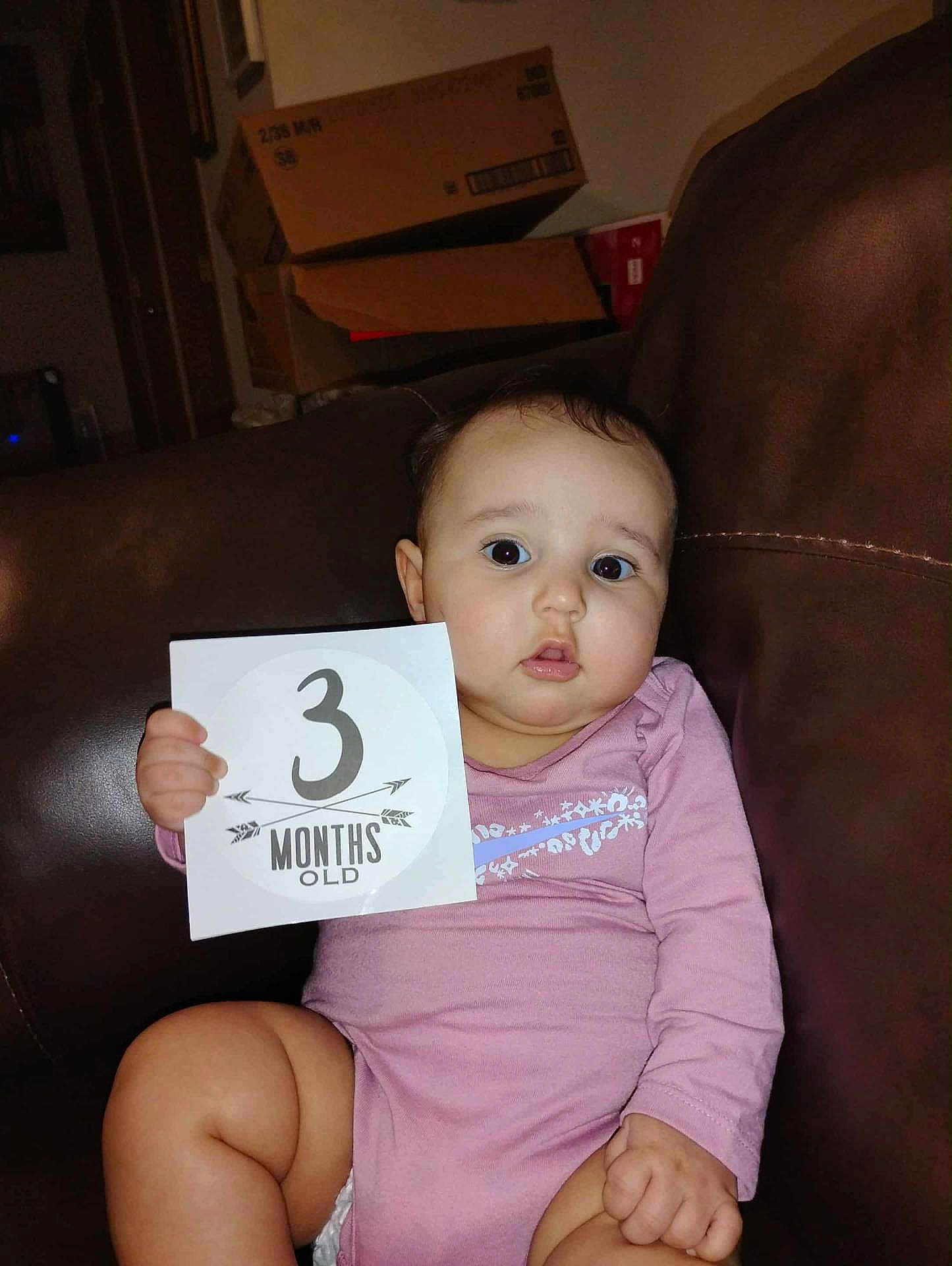 Jesy is registered to the contest to win money with this photo: 3_months_old, baby, cardboard_boxes, casual, child, couch, cute, expression, face, holding, home, indoor, infant, leather, person, pink_clothing, portrait, sign, sitting, young