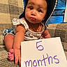 baby, infant, child, hat, blue_hat, floral_outfit, couch, patterned_couch, sign, hand, foot, face, expression, indoor, sitting, cute, portrait, young_child, person, purple_text