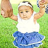 baby, child, grass, holding_hands, outdoor, skirt, white_top, headband, bow, earrings, two_adults, support, greenery, sunlight, cute, curious, standing, people, hands, nature