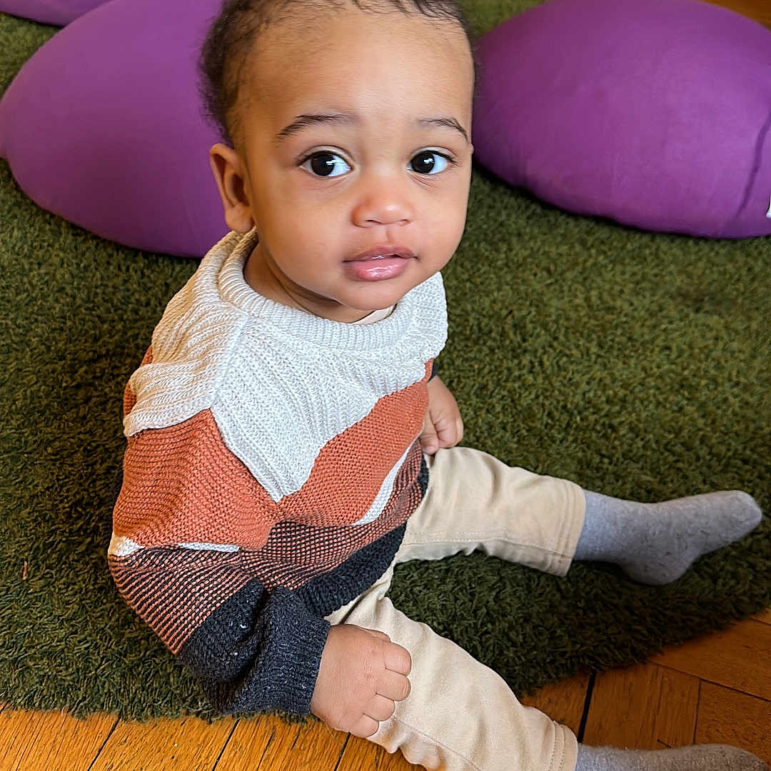 Noah Ryan participe au concours pour gagner de l'argent avec cette photo : big_eyes, casual_clothing, child, cozy, curious_expression, cute, flooring, green_rug, grey_socks, indoor, knitwear, playroom, portrait, purple_cushions, sitting, small_hands, sweater, toddler, wooden_floor, young_child