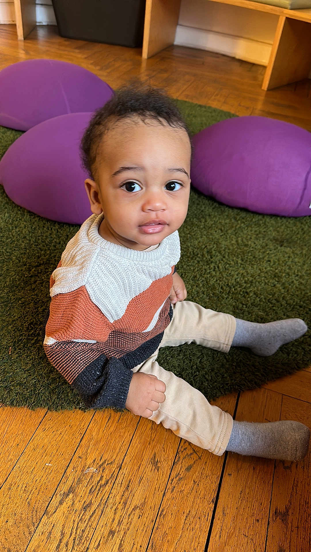Noah Ryan participe au concours pour gagner de l'argent avec cette photo : toddler, child, sitting, sweater, knitwear, grey_socks, green_rug, wooden_floor, purple_cushions, indoor, curious_expression, young_child, flooring, playroom, cozy, portrait, casual_clothing, small_hands, big_eyes, cute
