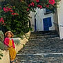 child, hat, smiling, sitting, cobblestone, pathway, flowers, pink_flowers, greenery, plants, stone_wall, stairs, building, white_wall, blue_shutters, outdoor, sunlight, happy, colorful_clothing, nature