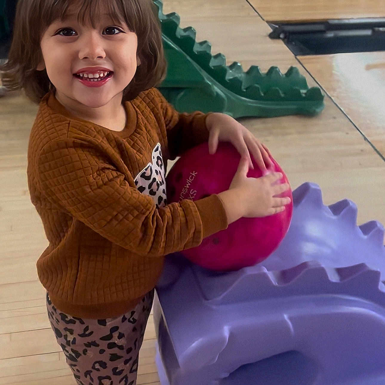 Zoey-Ann is registered to the contest to win money with this photo: activity, bowling_alley, bowling_ball, brown_hair, child, cute, fun, green_ramp, happy, indoor, leggings, leisure, person, playful, purple_ramp, recreation, smiling, sweater, toy, wooden_floor