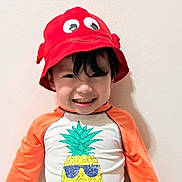 John is registered to the contest to win money with this photo: child, smiling, red_hat, pineapple, graphic_tshirt, orange_sleeves, happy, standing, indoor, young_child, cute, portrait, headwear, playful, casual_clothing, person, funny_hat, colorful, toddler, wall