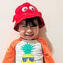 child, smiling, red_hat, pineapple, graphic_tshirt, orange_sleeves, happy, standing, indoor, young_child, cute, portrait, headwear, playful, casual_clothing, person, funny_hat, colorful, toddler, wall