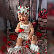 Millie is registered to the contest to win money with this photo: baby, child, smiling, white_clothing, headband, bow, strawberry, toy, sitting, decorations, fabric, cute, happy, indoor, portrait, sock, playful, wood_background, red, green