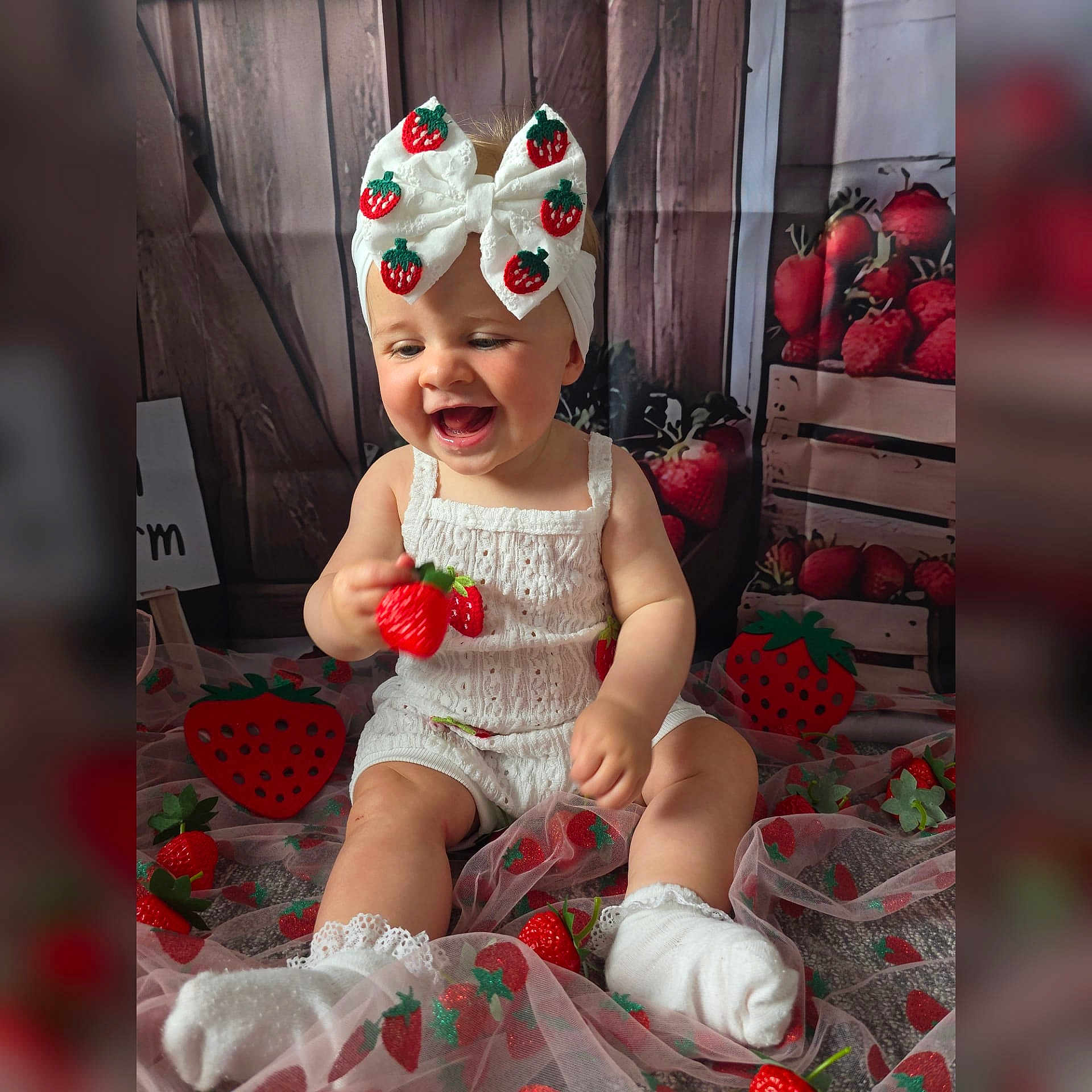 Millie is registered to the contest to win money with this photo: baby, child, smiling, white_clothing, headband, bow, strawberry, toy, sitting, decorations, fabric, cute, happy, indoor, portrait, sock, playful, wood_background, red, green
