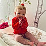 child, baby, toddler, smiling, red_shirt, hair_bow, bed, changing_pad, blanket, wall_mural, birch_trees, nursery, decor, hands_clapping, sitting, toes, pants, indoor, cute, portrait